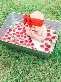 a baby sitting in a tub filled with water and strawberries on the grass, wearing a red headband