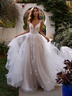 a woman in a wedding dress is standing on the sidewalk near some plants and flowers