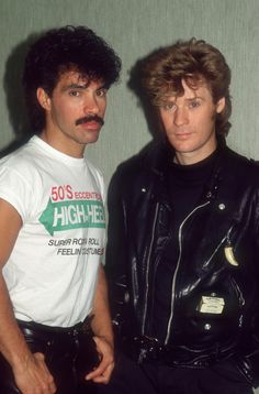 two men standing next to each other in front of a green wall wearing leather jackets and moustaches