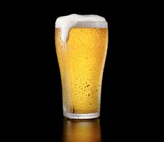 a tall glass filled with beer on top of a wooden table in front of a black background