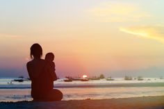 a woman and child sitting on the beach at sunset