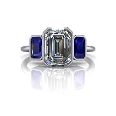 Three Stone Sapphire and Moissanite engagement ring, bezel set and featuring a thin band.*GEMSTONES: 8x6mm, 1.75 ct emerald cut colorless moissanite, DEF Color, VVS Clarity. (2) 5x3mm .40 ct, emerald cut blue sapphires.*Band width 1.5mm*2.75 carat total weight.*Our Moissanite is purchased in the USA. Stone arrives with a warranty card/certification of authenticity. Online registration is recommended.*The ring is custom made just for you in your ring size and metal preference. It is offered in so Engagement Ring Bezel, Bezel Engagement Ring, Three Stone Engagement Ring, Ring Bezel, Sapphire Band, Emerald Cut Rings, Online Registration, Forever One Moissanite, Blue Stones