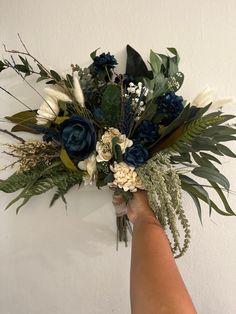 a hand holding a bouquet of flowers against a white wall with greenery and foliage