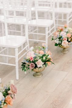 several white chairs are lined up with flower arrangements in gold vases on the floor