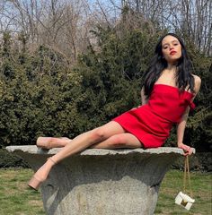 a woman in a red dress sitting on top of a stone bowl with her legs crossed