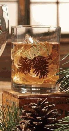 two glasses filled with liquid sitting on top of wooden boxes next to pine cones and evergreen needles