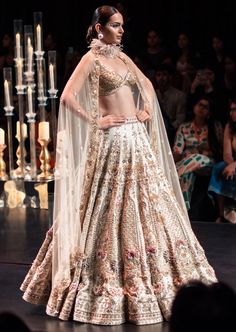 This enchanting earthy beige lehenga set is a beautiful blend of exuberance and grandeur. An ensemble that you will want this wedding season includes a lehenga, a blouse, a dupatta and a veil. Stitched to perfection, the lehenga features 3D embroideries with organza flowers using pearls, salli, cutdana, and stud tassels. The sleeveless blouse features a scintillating sweetheart neckline with handcrafted finish and a back hook closure. Paired with a heavy net dupatta, the cut-work detailing and intricate embroidery take center-stage. The lehenga comes with an attached cancan and the blouse has padding. Completed with a veil, this opulent ensemble silhouette will glimmer in the spotlight. Dry-clean only if required. Slight variation in color is possible due to digital photography. Anarkali Style Hand Embellished Sharara For Ceremony, Anarkali Ceremony Sets Hand Embellished, Traditional Beige Lehenga With Intricate Embroidery, Traditional Drape Lehenga With Intricate Embroidery, Beige Lehenga With Intricate Embroidery, Beige Lehenga With Sheer Dupatta Traditional Drape, Beige Lehenga With Sheer Dupatta For Diwali, Traditional Drape Beige Embroidered Lehenga, Traditional Hand Embellished Sharara For Ceremony