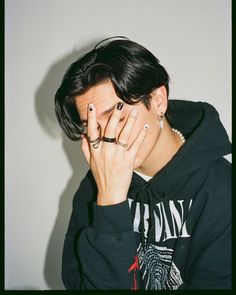 a young man covers his face with his hands while sitting in front of a white wall