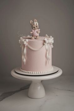 a pink cake decorated with an animal on top and ribbon around the edges, sitting on a white pedestal