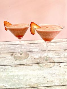 two martinis with strawberries in them on a wooden table next to a pink wall