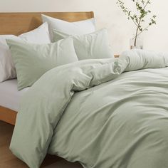 an unmade bed with light green sheets and pillows in a white room, next to a plant