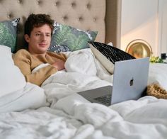 a young man laying in bed with his laptop on his lap and looking at the camera