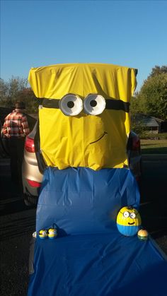 an inflatable yellow and blue object with eyes on it's head sitting next to a car