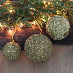 three gold ornaments hanging from a tree with lights in the background on a wooden surface