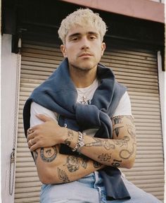 a man with tattoos on his arms sitting in front of a garage door and looking at the camera