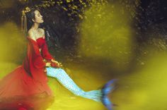 a woman in a red dress sitting on top of a mermaid tail while wearing a tiara