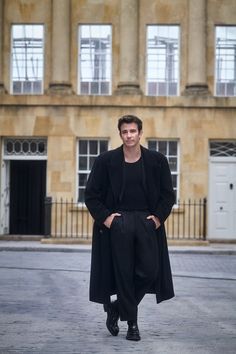 a man standing in front of a building wearing a long black coat and pants with his hands on his hips