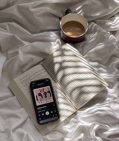 an open book, cup of coffee and cell phone on a bed with white sheets