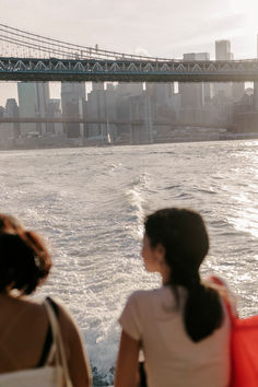 two women looking out at the water and bridge in the background with boats passing by