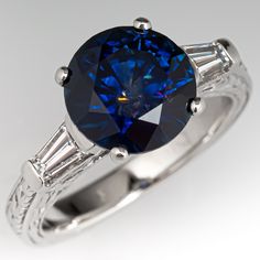 This stunning ring is centered with one (1) round brilliant cut Montana sapphire weighing 3.50 carats and set into a four-prong setting. The shoulders of the ring are each accented with one (1), channel set, tapered baguette cut diamonds. Engraved wheat motifs accented with ring. The ring measures 9.1mm at the top, rises 9.0mm above the finger, tapering to 3.0mm wide and 1.6mm thick at the base of the shank. This ring is currently a size 6. The sapphire shows a very visible natural crystal inclu Montana Sapphire Engagement, Montana Sapphire Engagement Ring, Diamond Sapphire Engagement Ring, Sapphire Diamond Engagement, Blue Engagement Ring, Blue Gemstone Rings, Sapphire Engagement Ring Blue, Golden Ring, Montana Sapphire