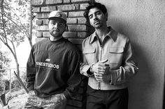 two men standing next to each other in front of a brick wall with trees behind them