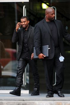 two men in black suits are talking on their cell phones and one is holding a folder