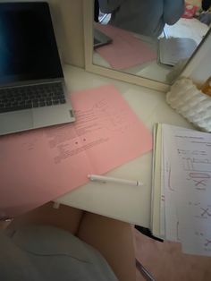 an open laptop computer sitting on top of a desk next to pink papers and a mirror