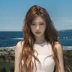 a young woman with long hair standing in front of the ocean and looking at the camera