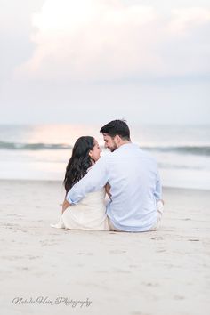 a man and woman sitting on top of a sandy beach next to the ocean at sunset