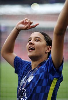 a woman in a blue shirt holds her hands up to the sky while standing on a soccer field