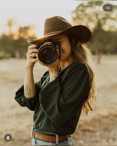 a woman wearing a hat and holding a camera up to her face while standing in a field