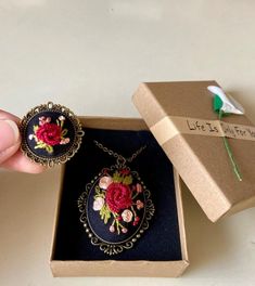 a hand holding a necklace with flowers on it in a box next to a flower brooch