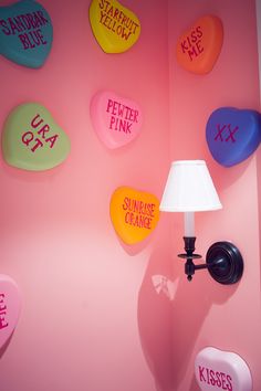 a pink wall with several different colored hearts on it and a lamp in the corner