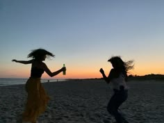 two women are running on the beach at sunset with their arms in the air and one is holding a bottle