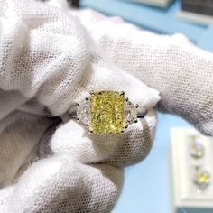 a fancy yellow diamond ring sitting on top of a white gloved hand in front of some jewelry