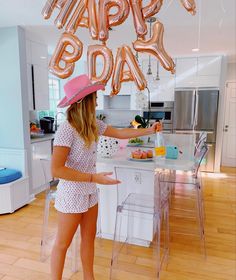 a woman wearing a pink hat standing in a kitchen with balloons that say happy birthday