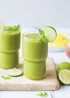 two glasses filled with green smoothie on top of a cutting board next to sliced limes