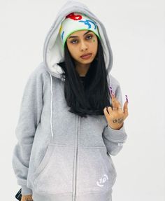 a woman with long black hair wearing a gray hoodie and holding her hand up