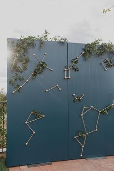 an art installation made out of sticks and vines on a blue wall in front of a brick walkway