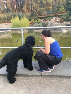 a woman kneeling down next to a black dog on top of a cement slab near a pond