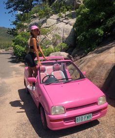 a woman standing on top of a pink car in the middle of a dirt road