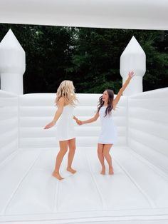 two women in white dresses holding hands and standing on an inflatable structure with trees in the background