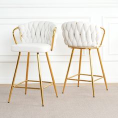 two white upholstered barstools with gold legs in front of a white wall