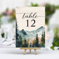 an easel with a table number on it in front of a mountain scene and flowers