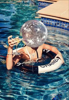 a woman floating in the water with a disco ball on her head and holding a drink