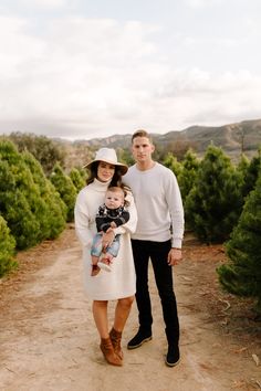 a man and woman standing on a dirt road holding a baby in their arms, surrounded by trees