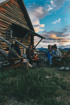 group of people sitting around a fire pit in front of a log cabin at sunset