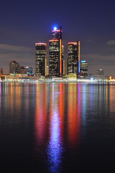 the city skyline is lit up at night with bright lights reflecting off the water in front of it