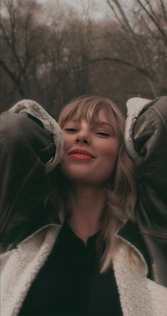 a woman with her hands on her ears and eyes closed, standing in front of trees