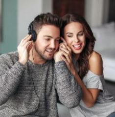 a man and woman sitting on a couch with headphones to their ears, smiling at each other